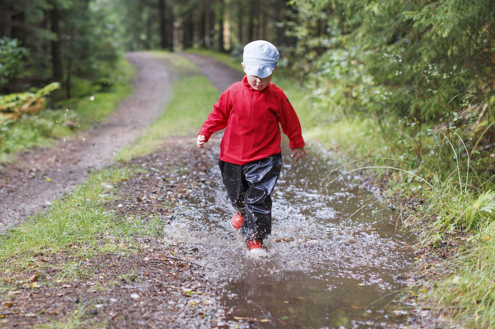 Liten pojke med keps och röd jacka vid vattenpöl i skogen.